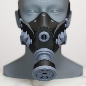 Peut inclure: Un masque à gaz noir et gris au design futuriste, avec un filtre proéminent et des pointes. Le masque est présenté sur une tête de mannequin grise. Le texte "NZL WORKS." est visible sur la tête.