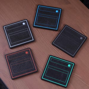 May include: Five black square coasters with colorful outlines and dot accents. Each coaster has a unique border color: white, blue, gray, red, and green. The coasters are arranged on a wooden surface.
