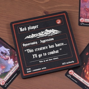 May include: A black coaster with red trim and white text that reads "Red player" and a quote about combat. Several Magic: The Gathering "Mountain" cards are scattered around the coaster on a wooden surface.