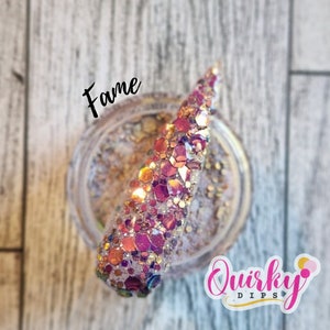 May include: A close-up of a small jar of iridescent glitter with a pink, gold, and purple color scheme. The glitter is labeled "Fame" and is from the brand "Quirky Dips".