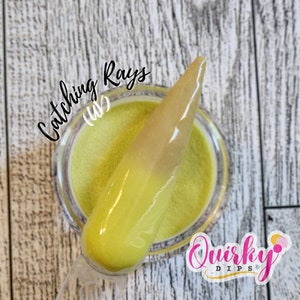 May include: A close-up of a clear jar filled with yellow and gold glitter nail dip powder. The jar is labeled "Catching Rays (UV)" and "Quirky Dips".