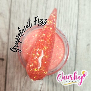 May include: A close-up of a small jar of pink glitter dip powder with a scoop of the powder on top. The jar has a white label with the text "Quirky Dips" and the text "Grapefruit Fizz" is written in black on the top of the jar.