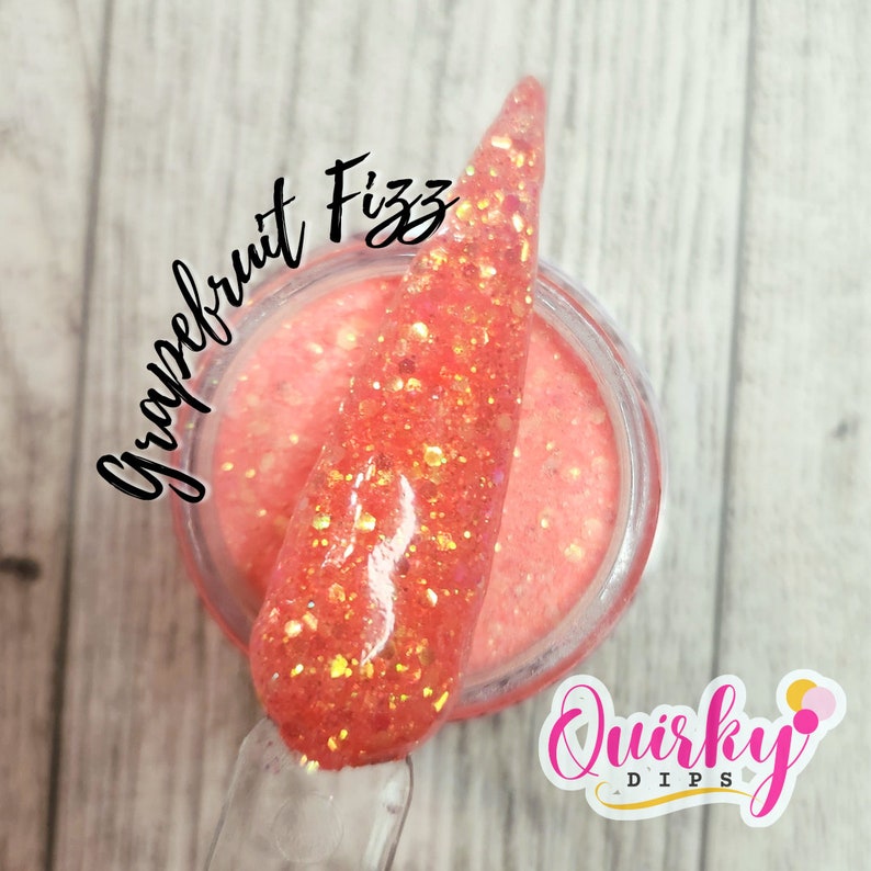 May include: A close-up of a small jar of pink glitter dip powder with a scoop of the powder on top. The jar has a white label with the text "Quirky Dips" and the text "Grapefruit Fizz" is written in black on the top of the jar.