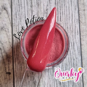 Puede incluir: Primer plano de un polvo para inmersión de uñas de color rojo con un acabado brillante. El polvo está en un frasco transparente con una etiqueta blanca que dice "Love Potion". El frasco está sobre una superficie de madera. El texto "Quirky Dips" es visible en la esquina inferior derecha de la imagen.