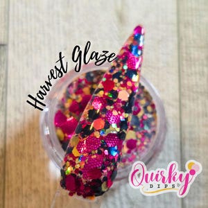 Op de afbeelding: Een doorzichtige pot met nagel-dip poeder met een monster op een doorzichtige stok. Het monster is bedekt met een mix van roze, zwarte, oranje en blauwe glitter. De woorden "Harvest Glaze" zijn in zwart cursief geschreven.