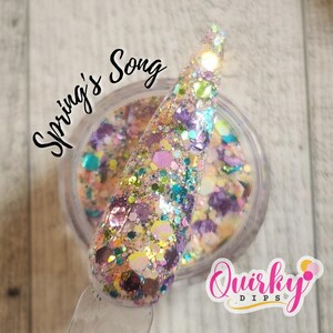 May include: A close-up of a clear jar filled with iridescent glitter. The glitter is a mix of light blue, pink, purple, and gold. The jar is labelled "Spring's Song" and "Quirky Dips".