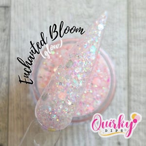 May include: A clear nail with iridescent glitter is held over a jar of pink glitter. The jar is labeled "Enchanted Bloom (glow)" and the logo "Quirky Dips" is in the bottom right corner. The background is a light wood grain.
