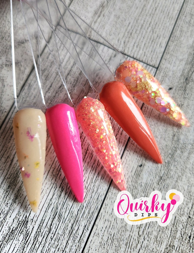 May include: Five different colors of nail dip powder displayed on clear plastic sticks. The colors are beige with gold glitter, hot pink, coral with gold glitter, orange, and a clear glitter with gold and iridescent glitter. The colors are arranged in a fan shape. The text "Quirky Dips" is visible in the lower right corner of the image.