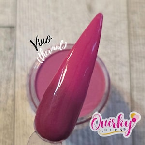 May include: A close-up of a nail dipped in a thermal powder, transitioning from a deep burgundy to a vibrant pink. The jar of powder is visible in the background. Text on the image reads "Vino thermal" and "Quirky Dips".