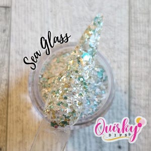 May include: A clear jar of nail dip powder with a clear applicator. The powder is a mix of iridescent glitter in shades of silver, teal, and gold. The words "Sea Glass" are written in black script. The logo for "Quirky Dips" is in the bottom right corner.