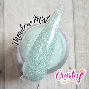 May include: A close-up of a clear container filled with a light blue glitter powder. The powder is on a clear applicator. The text "Meadow Mist" is written in cursive. The logo "Quirky Dips" is in the bottom right corner.
