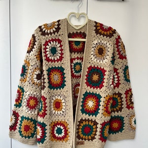 May include: A crocheted cardigan with a granny square pattern in beige, red, green, brown, and yellow. The cardigan has a V-neck and long sleeves.