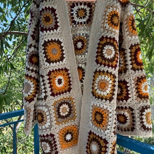 May include: A beige, open-front crochet cardigan with a floral pattern. The squares feature a mix of colors, including brown, orange, and cream. The cardigan is hanging on a pink hanger against a backdrop of green foliage.
