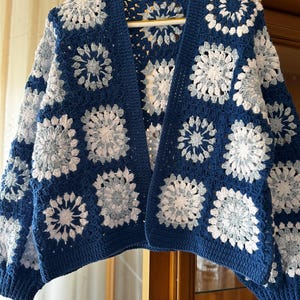 May include: A blue and white crocheted cardigan with a granny square pattern. The cardigan has a V-neck and long sleeves.