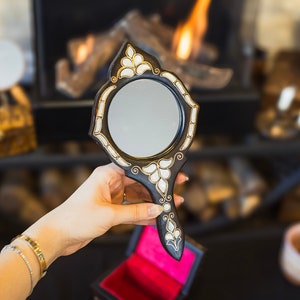 May include: A black hand mirror with intricate white and gold floral designs. The mirror has a round reflective surface and a long handle.
