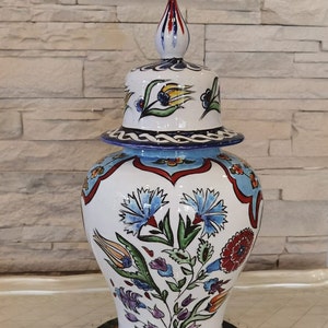 May include: A white ceramic vase with a blue and red floral design. The vase has a lid with a pointed top and a floral design. The vase is decorated with a variety of flowers, including tulips, carnations, and roses.