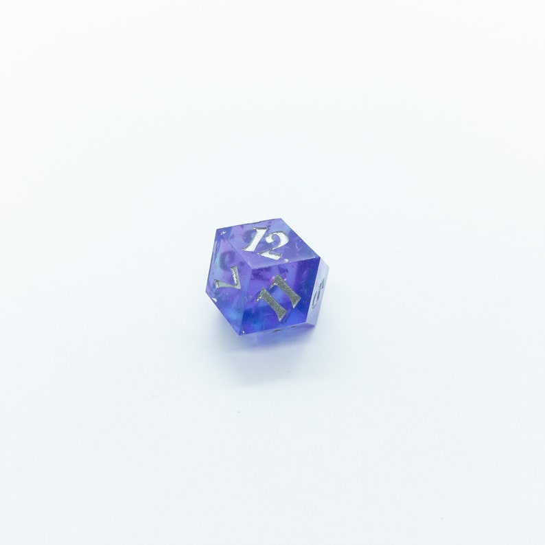 May include: A purple and blue iridescent d12 dice with silver numbers. The dice is showing the number 12.