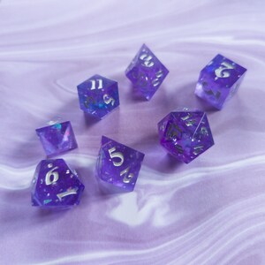 May include: A set of seven purple dice with silver numbers and glitter embedded in the plastic. The dice are on a purple and white background.