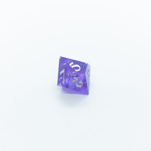 May include: A purple, iridescent, six-sided dice with silver numbers. The dice is showing the number 5.