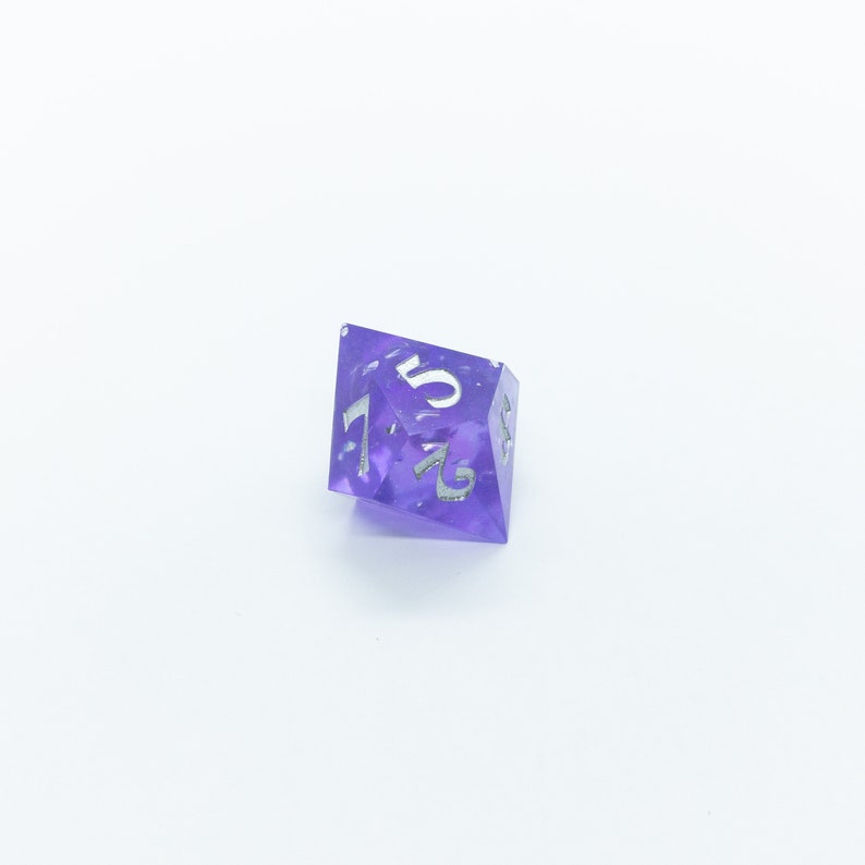 May include: A purple, iridescent, six-sided dice with silver numbers. The dice is showing the number 5.