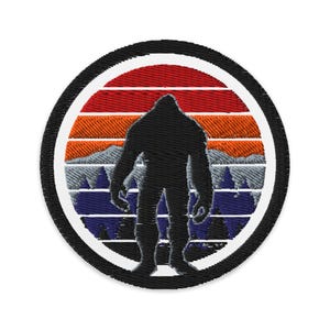Puede incluir: Parche bordado con la silueta negra de un Bigfoot sobre un fondo de puesta de sol. El diseño incluye rayas horizontales rojas, naranjas y azules, con una cordillera y árboles. El parche tiene un borde negro.