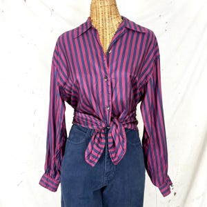May include: A button-down shirt with vertical stripes in burgundy and navy blue. The long-sleeved shirt is tied at the waist, and is paired with dark blue denim jeans. The shirt is displayed on a mannequin.