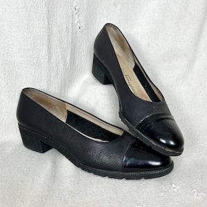May include: A pair of black leather pumps with a textured upper and a glossy patent leather toe cap. The shoes have a low, block heel and a rubber sole. The brand name "Ferragamo" is visible on the inner sole.