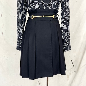 May include: A black pleated skirt with a high waist and a gold-tone belt buckle. The skirt is paired with a black and white patterned blouse. The skirt has a classic, tailored design.
