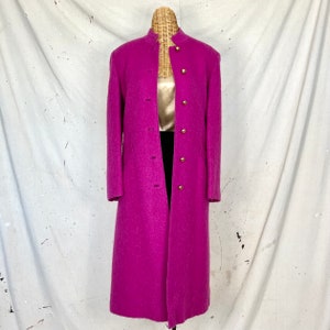 Puede incluir: Un abrigo largo de lana fucsia con cuello mandarín y botones dorados.
