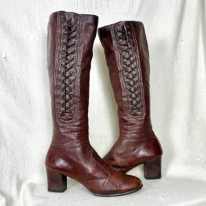 May include: A pair of knee-high brown leather boots. The boots feature a lace-up design on the front and a block heel. The leather has a textured appearance, and the boots are set against a neutral background.