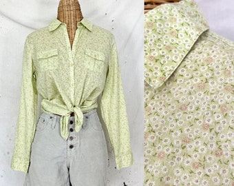 Puede incluir: Una camisa verde claro con botones y estampado floral. La camisa tiene mangas largas y dos bolsillos en el pecho. La camisa está anudada a la cintura y se lleva con pantalones cortos grises claros.