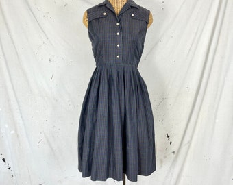 Könnte beinhalten: Ein ärmelloses kariertes Kleid mit Knopfleiste und einem gerafften Rock. Das Kleid ist dunkelblau mit einem dezenten grün-weiß karierten Muster.