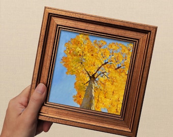 May include: A framed painting of a tree with yellow leaves against a blue sky. The artwork is in a brown wooden frame. The painting is held by a hand.
