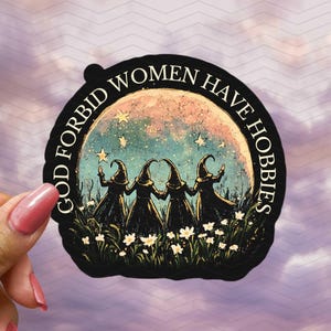 May include: A black sticker featuring the text "GOD FORBID WOMEN HAVE HOBBIES" around an illustration of four witches in black robes and hats, set against a moon and stars. White flowers are at the base of the image.