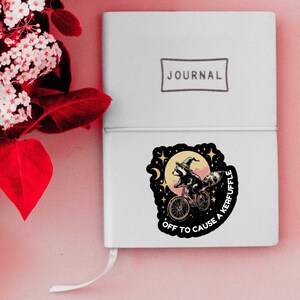 Puede incluir: Un diario blanco con la palabra "JOURNAL" impresa en la portada. Una pegatina de un mapache montando en bicicleta con un sombrero de bruja est&aacute; en el diario. La pegatina dice "OFF TO CAUSE A KERFUFFLE".