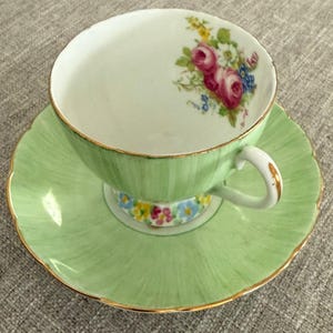 May include: A green teacup and saucer set with a floral design. The cup and saucer have a gold trim. The floral design features pink roses and other flowers.