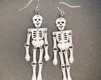 May include: A pair of skeleton earrings with a white finish. The earrings are made of plastic and have a dangling design. Each earring features a skeleton with a smiling face and articulated limbs.