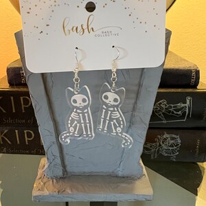 May include: A pair of clear acrylic earrings featuring a skeleton cat design. The earrings are hanging from silver hooks and are packaged on a white card with the text "bash collective" printed on it.