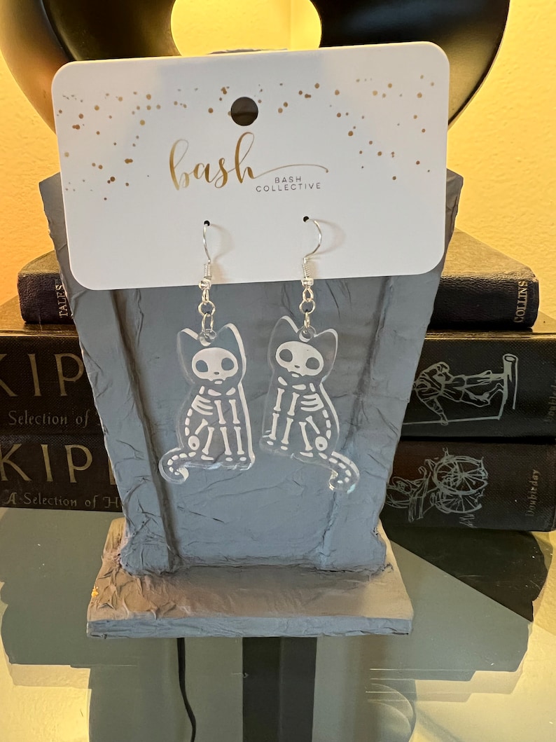 May include: A pair of clear acrylic earrings featuring a skeleton cat design. The earrings are hanging from silver hooks and are packaged on a white card with the text "bash collective" printed on it.