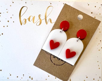 May include: White arch-shaped earrings with red heart charms and red tops. The earrings are displayed on a brown card with the text "bash collective" printed on it.
