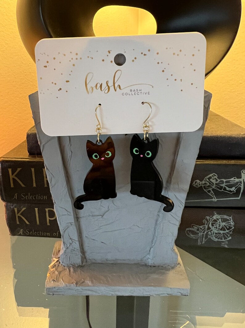 May include: A pair of black cat earrings with silver hooks. The earrings are on a white card with the text "bash collective" printed on it.