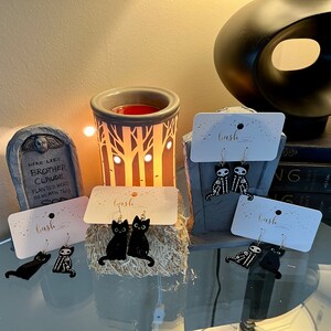 May include: Three pairs of Halloween-themed earrings featuring black cats and skeletons. The earrings are displayed on white cards with the word "bash" printed on them.