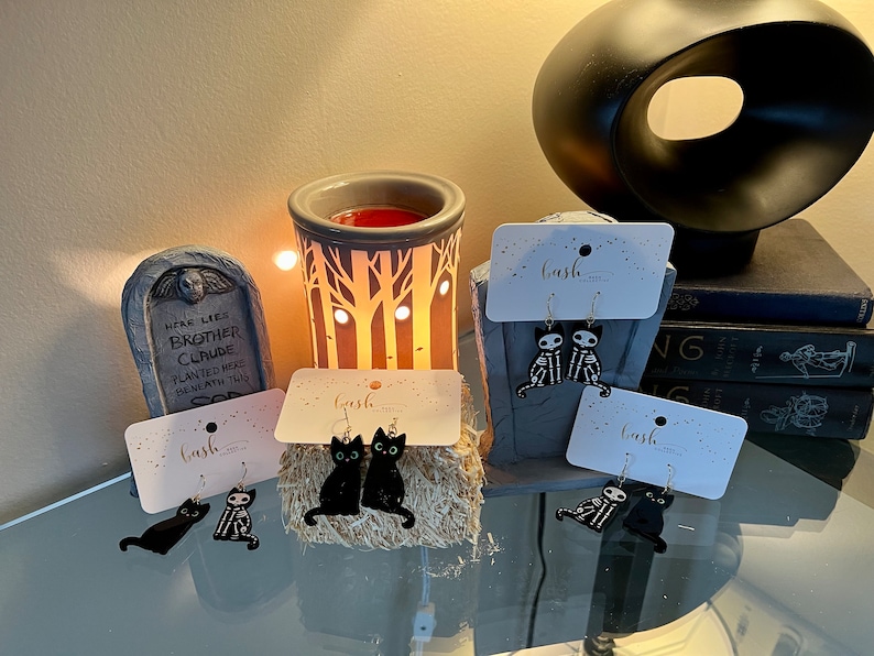 May include: Three pairs of Halloween-themed earrings featuring black cats and skeletons. The earrings are displayed on white cards with the word "bash" printed on them.