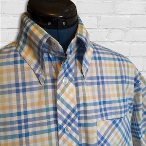 Könnte beinhalten: Ein Button-Down-Hemd mit einem Karomuster in Blau-, Gelb- und Weißtönen. Das Hemd hat einen Button-Down-Kragen und eine Brusttasche. Das Hemd wird auf einer Schaufensterpuppe präsentiert.