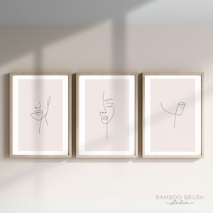 May include: Three framed minimalist line art prints of faces with a blush pink background. The prints are in wooden frames and are hung on a light beige wall.