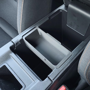 May include: A black car interior with a center console that has two storage compartments. The compartments are open and empty. The console is made of black plastic and has a textured surface.