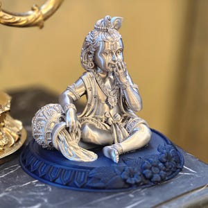 Könnte beinhalten: Silberfarbene Statue einer sitzenden Figur, geschmückt mit aufwendigem Schmuck und einer Krone. Die Figur ruht auf einem blauen Sockel mit floralen Details. Ein dekoratives Objekt ist im Hintergrund sichtbar.