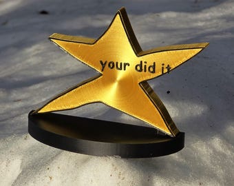 May include: A gold star-shaped award with the words "your did it" engraved on it. The star is sitting on a black base.