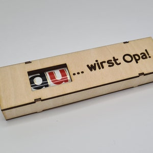 Puede incluir: Una caja de madera con una ventana recortada que revela las letras "du" y el texto "... wirst Opa!" grabado en la parte superior.
