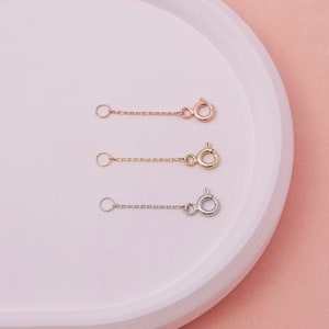 May include: Three necklace extenders in rose gold, gold, and silver. Each extender features a small chain with a spring ring clasp on one end and a small ring on the other. The extenders are displayed on a white, curved surface against a pink background.